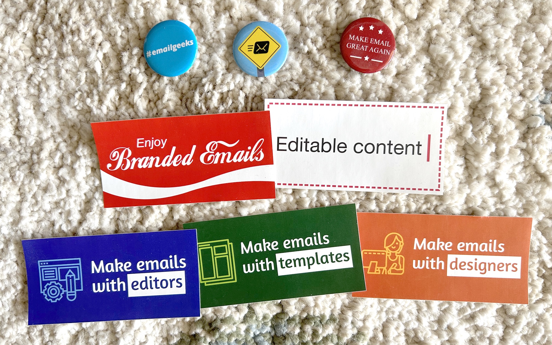 A photo of stickers and pins made for #emailgeeks that say: 'Enjoy Branded Emails', 'Editable content', Make email great again', '#emailgeeks', 'Make emails with editors', 'Make emails with templates', 'Make emails with designers'