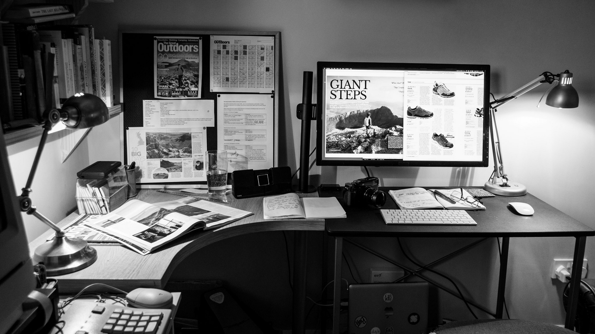 A black and white photo of an editorial magazine office desk, with a computer monitor, keyboard, and photo camera.