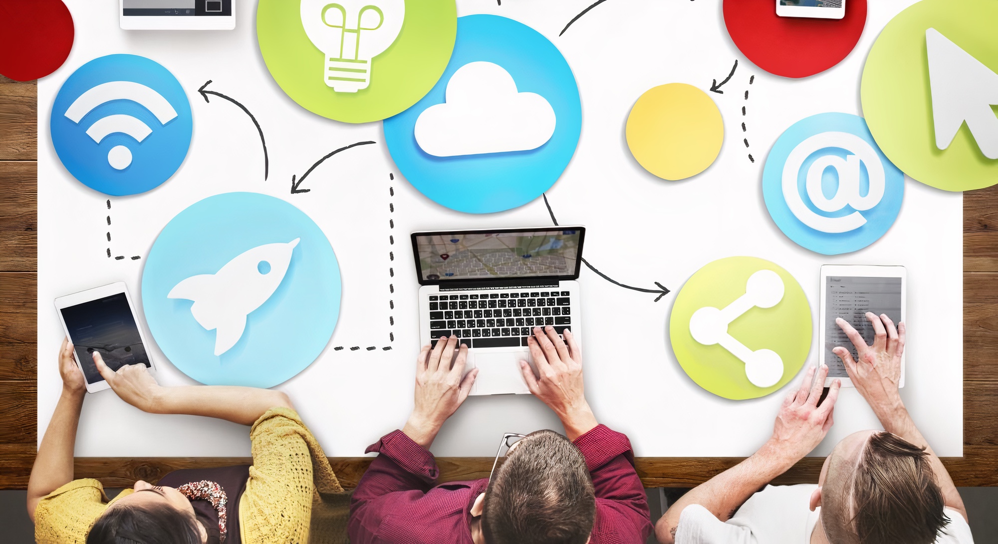An overhead shot of three team members working together on their devices with icons representing their tools laid out on the table in front of them.